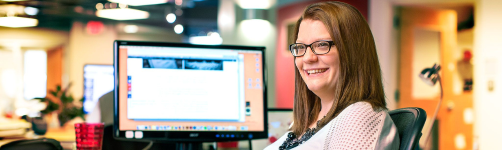 woman with glasses seated in front of computer screen in office, smiling widely